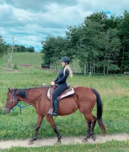 The side profile of a young Caucasian woman with blonde hair wearing all black, including black riding boots and a black helmet, riding a brown horse with black hair along a skinny dirt trail with a green field and green trees in the background