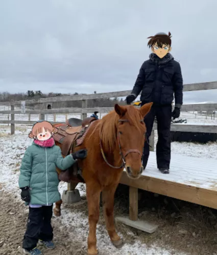 Two kids posing with a brown horse. One kid, a boy, is standing on the horse's left on top of a wooden platform wearing all black. A girl wearing a teal winter coat is standing on horse's right on the ground. It is winter ad there is snow on the ground.