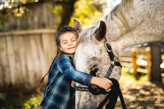 A young Caucasian girl wearing a blue plaid shirt hugging a white and grey horse with her eyes closed and a grin on her face. The horse is leaning down and into the girl and also has its eyes closed.